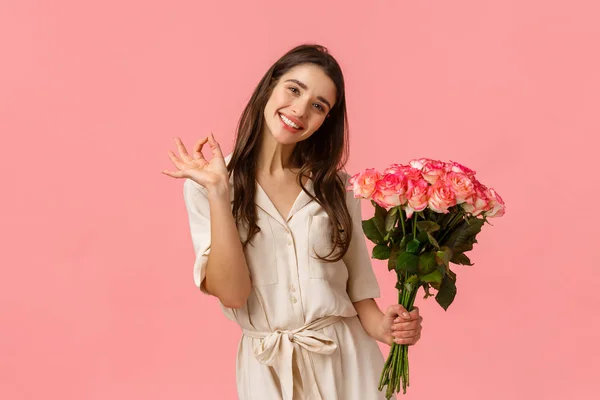 Preciosa, soñadora y coqueta morena hembra en vestido, cabeza inclinada y sonriente encantada, mostrando bien, buenas rosas de celebración, tiene regalo en el día de San Valentín, hermosas flores de ramo, fondo rosa — Foto de Stock