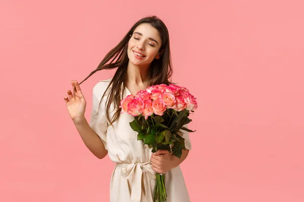 Concepto de ternura, deleite y día de San Valentín. Mujer morena encantadora, encantadora y sensual en vestido, enrollando rizos en el cabello felizmente cerrar los ojos y soñar, recibir entrega de flores, sostener rosas — Foto de Stock