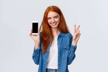 Hoş ve neşeli, kot gömlekli, barış jesti gösteren, gülümseyen ve akıllı telefon tutan kızıl saçlı bir kadın. Program uygulaması, şirket web sitesi ya da mobil ekranda alışveriş yapan internet mağazası, beyaz arka plan.