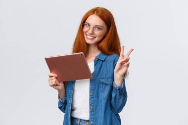İnsanlar, iş ve eğitim kavramı. Gözlüklü neşeli, çekici, kızıl saçlı kız öğrenci, uzak online kurslar çalışıyor, dijital tablette video izliyor, barış işareti gösteriyor ve gülümsüyor.