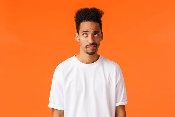 Indecisive and skeptical, gloomy african-american hipster guy with moustache, afro haircut, smirk and looking up unsure, having doubts, dont know how act, standing orange background — Stock Photo, Image