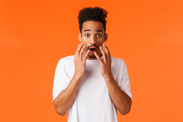 Scared, freaked-out and shocked african-american young hipster guy looking at something scary or frightening, standing concerned over orange background, hold hands near opened mouth — Stock Photo, Image