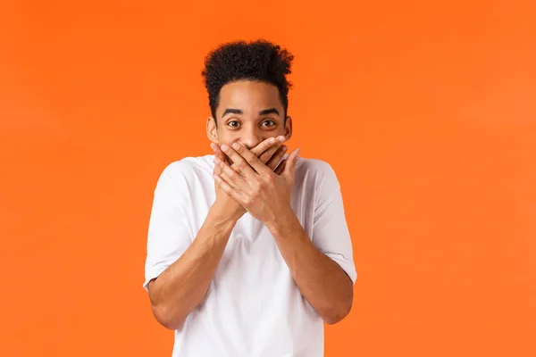 Guy made funny prank trying hold laugh. Hilarious and amused african-american male in white t-shirt, giggle cover mouth with palms and staring amazed camera, chuckling, standing orange background — Stock Photo, Image