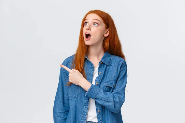 Tailliertes Porträt erstaunt, sprachlos hübsche rothaarige Teenager-Collage-Studentin, Ingwer langes Haar, keuchender Kiefer und fragender Blick in die linke obere Ecke, der auf faszinierendes Produkt zeigt — Stockfoto