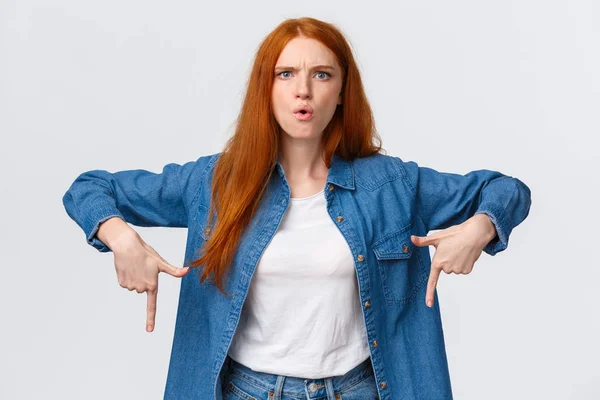 Explain this now. Serious-looking mad and perplexed, confused redhead woman pointing down, frowning and grimacing, dont unserstand who made all mess, feeling angry over white background — Stock Photo, Image