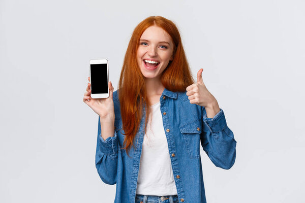 Girl showing friend cool new mobile game. Attractive cheerful redhead woman holding smartphone, introduce application, telephone app, make thumb-up and smiling in approval, recommending