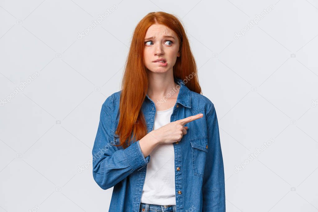 Awkward and nervous cute timid redhead teenage girl having fear or embarrassing thing say, pointing looking right anxious, biting lip and frowning, confessing in crime, feel guilty