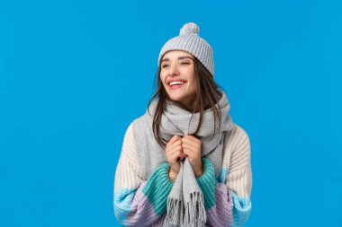 Kışlık şapkalı, atkılı ve süveterli neşeli esmer kadın, Noel tatilinin tadını çıkarıyor, karda oynuyor, mavi arka planda neşeli bir şekilde duruyor.