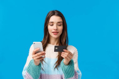 Kışın süveter giyen, belden yukarı portre tarzı modern bir kadın internetten sipariş veriyor, internetten alışveriş yapıyor, akıllı telefondan kredi kartı ekliyor, kredi kartı ve cep telefonu tutuyor, mavi arka plan kullanıyor.