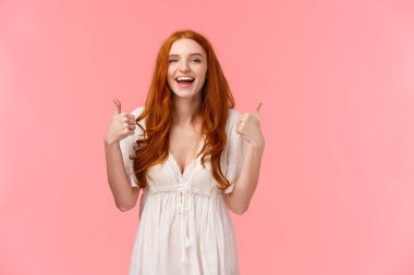 Belden yukarı, neşeli, neşeli, beyaz elbiseli kızıl saçlı kadın, başparmağını kaldırmış ve gülümsüyor, ürün gibi, tavsiye et, olumlu görüş bildir, pembe arka planda dur