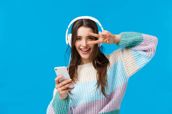 Happy sassy przystojny brunetka kobieta w słuchawki, sweter zimowy, korzystających z ulubionych piosenek w nowych słuchawek, show peace, disco gest trzymając smartfona, uśmiechnięty aparat zadowolony — Zdjęcie stockowe