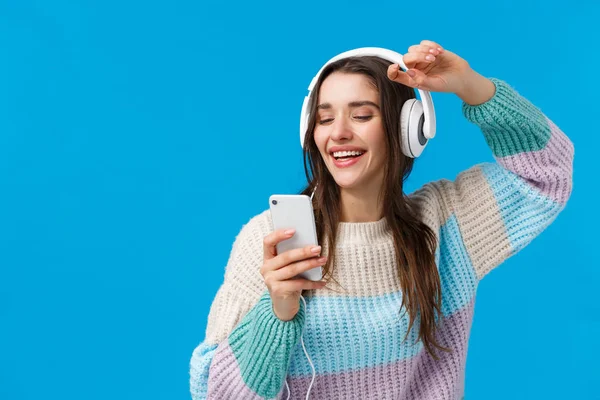 Glad karismatisk och bekymmerslös leende europeisk kvinna njuter av fantastisk musik kvalitet ljud i nya hörlurar som mottagits som julklapp, höja händerna upp hålla smartphone, blå bakgrund — Stockfoto