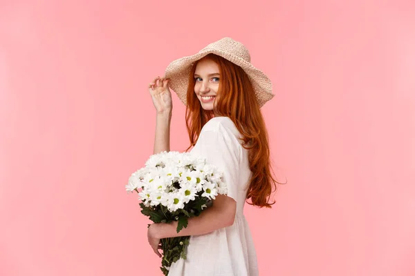 Romantyczna, głupia i czuła ruda kobieta w uroczym kapeluszu, sukience, trzymając bukiet białych kwiatów, aparat i uśmiechnięty coquettish, flirtując z chłopakiem na różowym tle — Zdjęcie stockowe