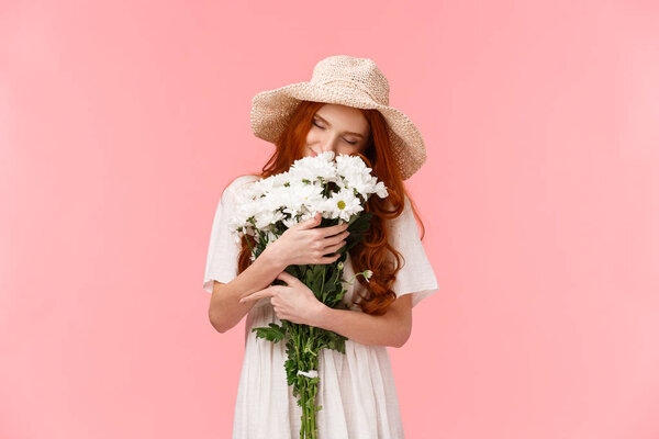 Romance, love and relationship concept. Romantic dreamy, alluring redhead girl sniffing flowers, close eyes and hugging beautiful bouquet, standing pink background