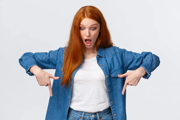 Waist-up portrait impressed, curious and astonished redhead cheerful girl in denim shirt, drop jaw, looking and pointing down starled speechless, standing white background amused — Stock Photo, Image