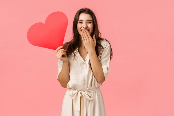 Concepto de felicidad, relación y romance. Atractiva mujer bastante femenina con cabello rizado oscuro, vestido, risueño cubrir la boca como risita, sosteniendo rojo lindo corazón de cartón, fondo rosa —  Fotos de Stock