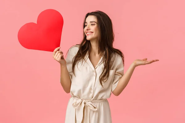 Sorpresas, relación y concepto romántico. Atractiva morena despreocupada y feliz sonriente en vestido, levantando las manos divertidas y alegres mirando a la izquierda, sosteniendo la tarjeta de amor del corazón, fondo rosa —  Fotos de Stock