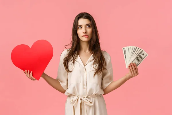 Romance, wealth and modern lifestyle concept. Alluring good-looking brunette female in dress, holding cash, money and love heart, biting lip look upper right corner unsure, making decision