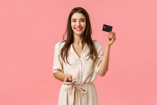 Publicidade, tecnologia e conceito de estilo de vida digital. Despreocupado atraente jovem mulher em vestido lindo mostrando cartão de crédito e segurando smartphone, sorrindo compra on-line, fundo rosa — Fotografia de Stock