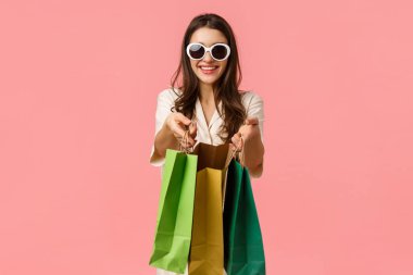 Alışveriş, tüketici konsepti. Neşeli, çekici esmer kız, alışveriş torbaları tutuyor ve gülümsüyor, gözlük takıyor, tezgahlarda geziniyor, pembe arka planda neşe içinde duruyor.