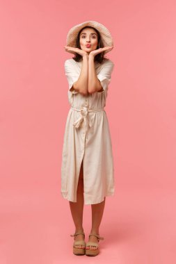 Tam boy dikey fotoğraf, dudaklarını kıvıran, dudak büken, öpüşen, yüze dokunan, kozmetik ürünleri tanıtan, güzel bir elbise ve şapka içinde duran, pembe arka plan pozu veren aptal kadın.