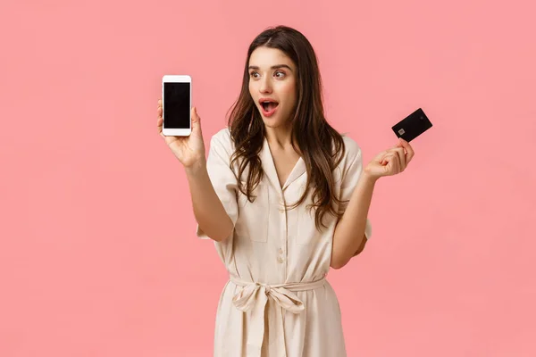 Забавная и удивленная, красивая белая женщина в платье, держащая кредитку и показывающая что-то потрясающее и классное на экране смартфона, стоящая розовый фон радостно — стоковое фото