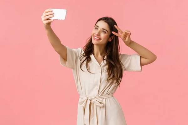 Carefree emotive, happy smiling brunette female in dress, holding smartphone, taking selfie and make peace sign, tilt head and smiling, sending positive vibes to followers, standing pink background — Stock Photo, Image
