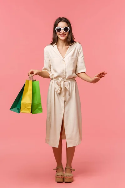 Ganzes vertikales Porträt elegante gut aussehende Frau im Urlaub, Einkaufen und Spaß in Einkaufszentren, trägt Sonnenbrille, leichtes Kleid, trägt Taschen aus dem Geschäft, steht rosa Hintergrund lächelnd — Stockfoto