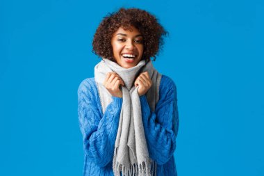 Hoş, karizmatik genç bir kadın kış kıyafetleri aldı, kendini sıcak tutmak için eşarba sardı, gülümsedi ve güldü rahatladı, mavi arka planda dikildi.