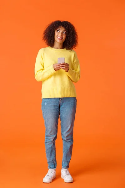 Full-length vertical shot dreamy cute african-american woman thinking-up what write, holding smartphone thinking looking up and smiling imaging things, standing orange background joyful