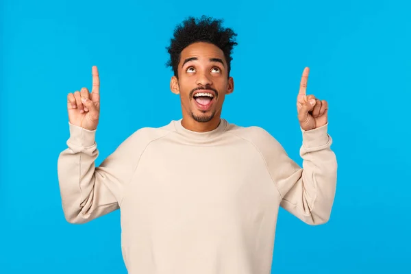 Un tipo que ve una señal interesante, repitiéndola desde arriba. Atractivo afroamericano masculino en suéter de invierno, apuntando hacia arriba y sonriendo impresionado, feliz encontrado excelente promo, fondo azul —  Fotos de Stock