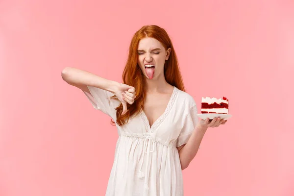 Ignorant, picky and snobbish redhead birthday girl hate b-day cake, holding plate with dessert, showing tongue grimacing and make thumb-down in disapproval, aversion and disgust — Stock Photo, Image