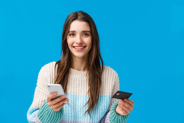 Concepto de pago, compras online y banca. Atractiva compra de mujer sonriente en la tienda de Internet, la celebración de la tarjeta de crédito y el teléfono inteligente, mirando a la cámara, la compra de regalos de vacaciones de invierno, fondo azul — Foto de Stock