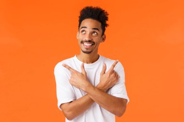 Hevesli, heyecanlı, bıyıklı Afro-Amerikan erkek, rüya gibi ve tasasız, mutlu bir şekilde gülümseyen, sağı solu işaret eden, seçim yapan, ne alacağına karar veren, turuncu arka planlı.