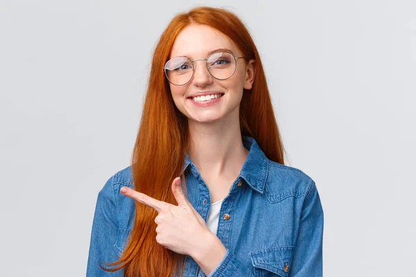 Κοντινό πλάνο πορτρέτο χαριτωμένο χαρούμενο κοκκινομάλλα, foxy κορίτσι σε γυαλιά, δείχνει μέρος για να κάνετε κλικ στην ιστοσελίδα, ακολουθήστε ή εγγραφείτε, δείχνοντας το δάχτυλο αριστερά και χαμογελώντας ευχάριστο, με φιλική στάση, λευκό φόντο — Φωτογραφία Αρχείου