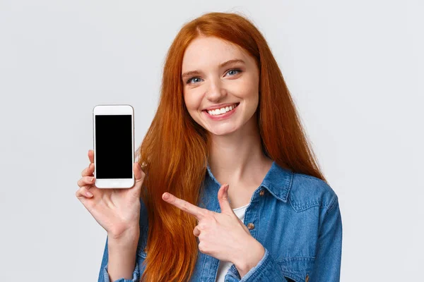 Dziewczyna znalazła swoją randkę w apce. Wesoła, atrakcyjna ruda kobieta z niebieskimi oczami daje rady, pokazując wyświetlacz smartfona i wskazując palcem ekran mobilny, uśmiechając się dać rekomendację — Zdjęcie stockowe