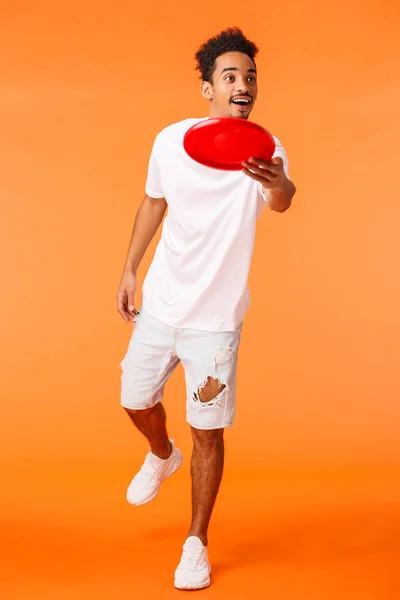 Full-length verticale shot zorgeloos knappe Afrikaans-Amerikaanse millennial guy genieten van de zomer vrije tijd, outdoor activiteit, gooien frisbee glimlachen en kijken naar vriend, oranje achtergrond — Stockfoto