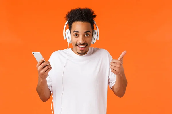 Portret w talii wesoły, charyzmatyczny, afrykański Amerykanin z wąsami, afro haircut, trzymający smartfona pokazującego kciuk w górę Słuchać muzyki w słuchawkach, uśmiechnięty, zadowolony świetną jakość dźwięku — Zdjęcie stockowe