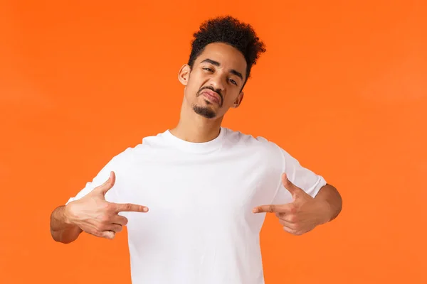 Sassy and confident assertive african-american man, acting cool and cheeky, pointing himself proud, boastful standing orange background, show-off, making impression, wearing white t-shirt — Stock Photo, Image