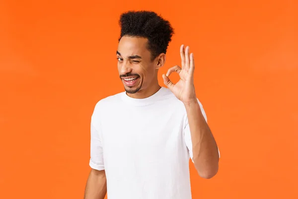 Rely on me. Outgoing relaxed and confident young attractive african-american male encourage, being assertive, showing okay sign and wink, reassure all is good, orange background — Stock Photo, Image