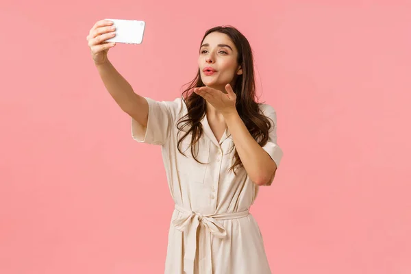 Härlig och anbud, lockande brunett hona i klänning innehav smartphone, ta selfie och blåser kyss på mobilkamera, ström live video för blogg, fotografering över rosa bakgrund — Stockfoto