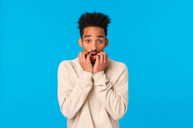 Korkmuş ve paniğe kapılmış genç Afrikalı Amerikalı korkmuş, korku filmi izleyen, parmaklarını ısıran ve yıldızlı kameraya bakan korku ve gerginliği ifade eden mavi arka plan.
