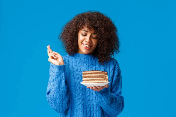 Cute and silly hopeful young african-american woman in sweater, with curly afro haircut, cross fingers good luck, praying and holding plate with delicious big piece cake, standing blue background