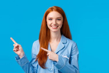 Gecelikli, mutlu, kızıl saçlı bir kadın, sol üst köşeyi işaret ediyor, dostça gülümsüyor, arkadaşının yer bulmasına yardım ediyor, mükemmel reklam fotokopisi çekiyor, ürün öneriyor, mavi arka planda duruyor.
