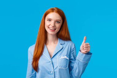 Kız tavsiye edilen ürünü onaylıyor. Neşeli, kızıl saçlı, pijama giymiş güzel bir kadın, uyumaya hazır, bilgiyi doğrulayan, evet işareti yapan, gülümseyen, mavi arka planda duran