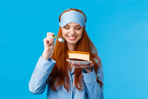 Waist-up portrait cheerful and delighted young caucasian redhead woman in pyjama and sleep mask, holding slice cake and spoon smiling delighted, looking at tasty dessert, wear nightwear