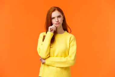 Şüpheci ve kararsız kızıl saçlı, ciddi görünümlü kadın zor kararlar alıyor, seçim hediyesi veriyor, kış tatilini geçiriyor, alışveriş koridorlarına bakıyor, dudaklarına dokunuyor, düşünüyor, turuncu arka plan düşünüyor.