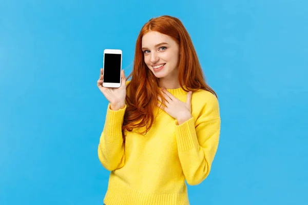 Impressed and fascinated redhead sassy woman touch heart and flirty gazing camera showing sexy guy photo she met dating app, holding smartphone with display facing camera, blue background — Stock Photo, Image