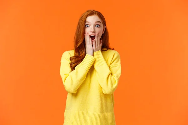 Impressed and speechless redhead woman found out awesome news, gasping, drop jaw astonished, touch cheeks and stare startled camera, christmas shopping season started, orange background