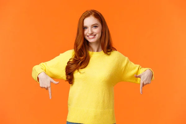 Precisas de ver isto, vê isto. Menina ruiva lindo alegre em suéter amarelo, sorrindo e apontando para baixo, recomendar o produto, publicidade compras férias vendas, fundo laranja — Fotografia de Stock